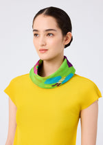 A woman poses against a plain light gray background wearing a yellow textured short-sleeve top and the FLOWER DOME PLEATS STOLE by me ISSEY MIYAKE, featuring vibrant botanical motifs in pleated green.