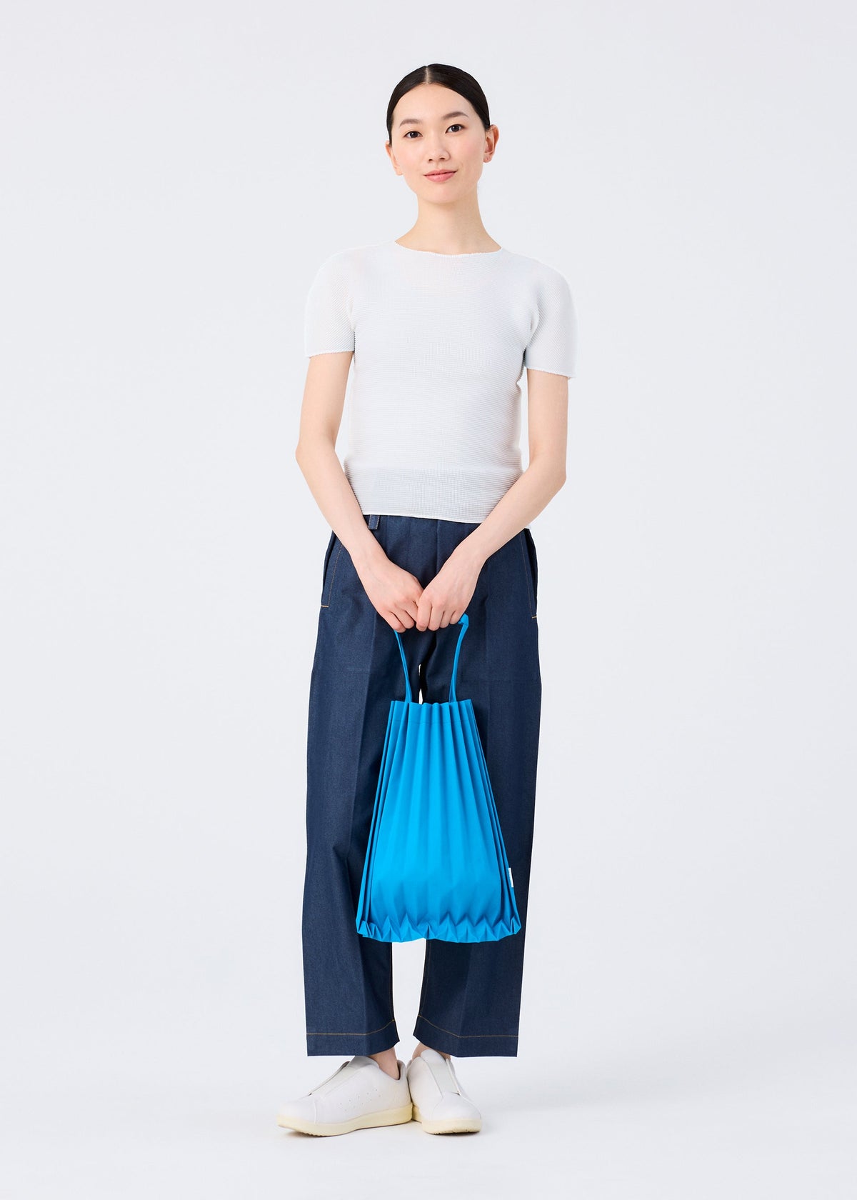 A person stands against a plain background wearing a white short-sleeve top, wide-leg blue pants, and white shoes, holding the vivid TRUNK PLEATS BAG 12 by me ISSEY MIYAKE with both hands in front of them. view 7 of 7