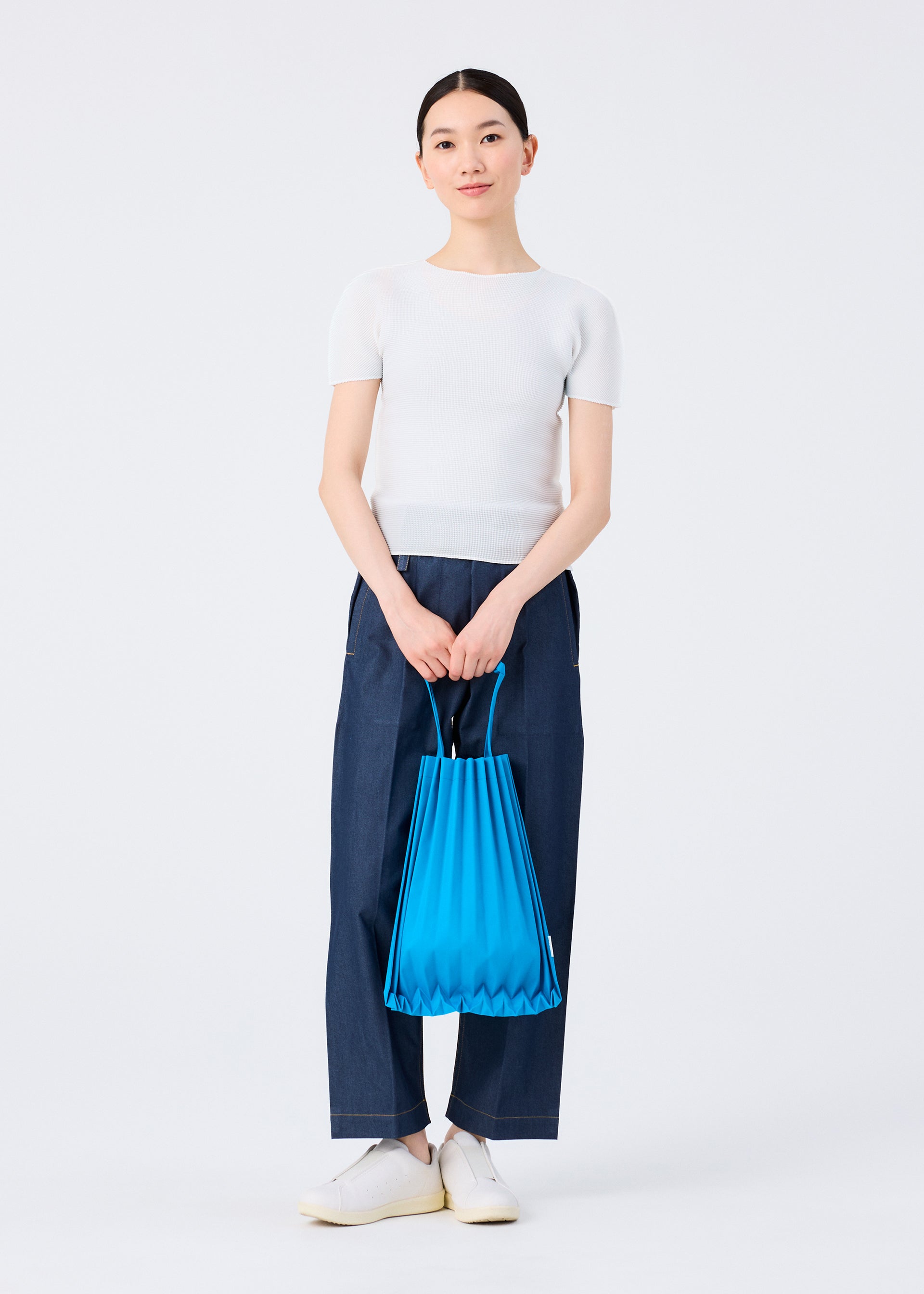 Zoomed view A person stands against a plain background wearing a white short-sleeve top, wide-leg blue pants, and white shoes, holding the vivid TRUNK PLEATS BAG 12 by me ISSEY MIYAKE with both hands in front of them.