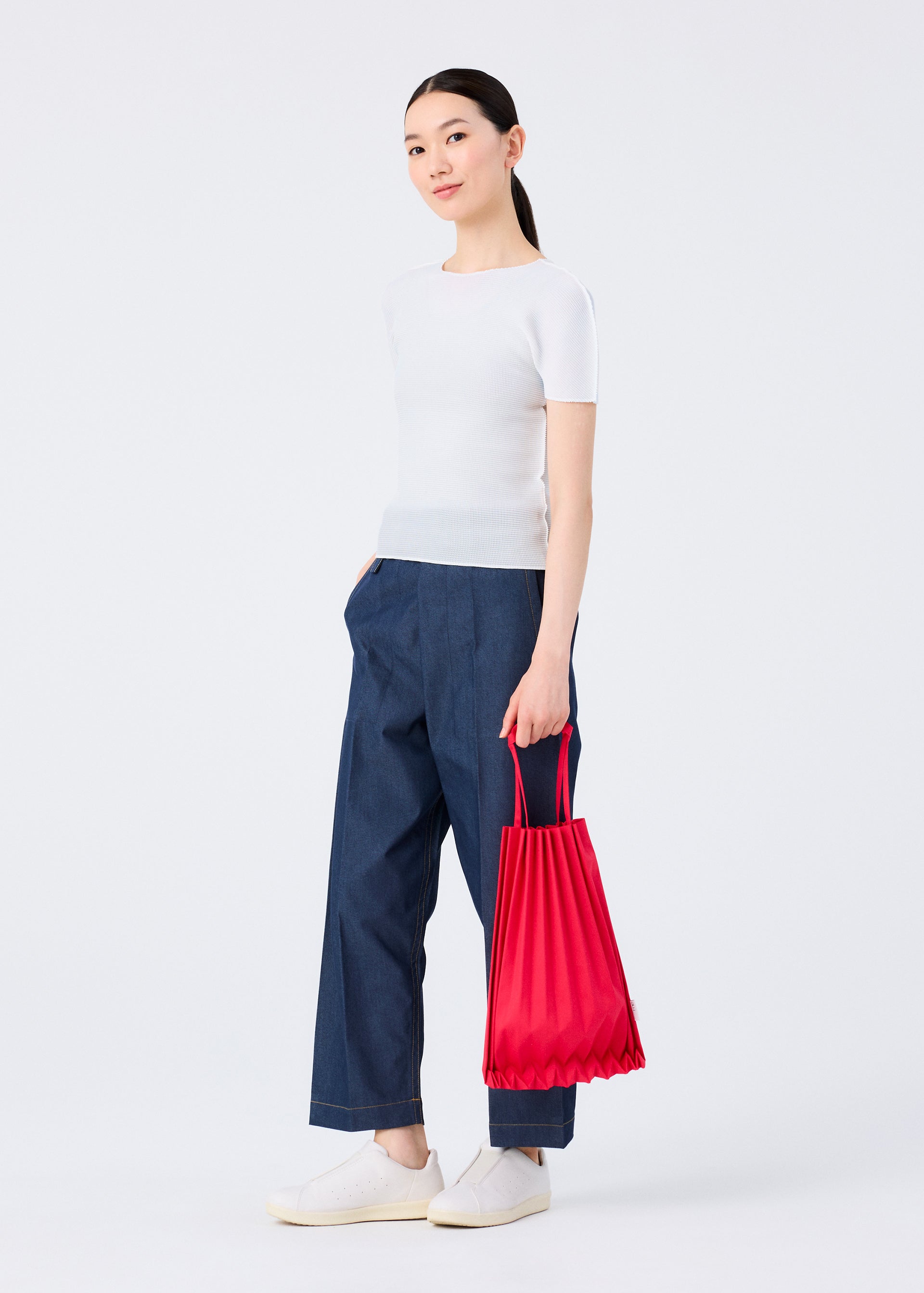 Zoomed view A woman with her hair tied back smiles softly, standing against a white background in a white short-sleeve top and wide blue pants, holding the vivid TRUNK PLEATS BAG 12 by me ISSEY MIYAKE, featuring its distinctive handle.