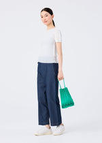 A person stands against a plain white background, wearing a fitted white short-sleeve top, loose blue denim pants, white slip-on shoes, and holding the TRUNK PLEATS BAG 12 by me ISSEY MIYAKE—a small green mini tote with vivid pleats.