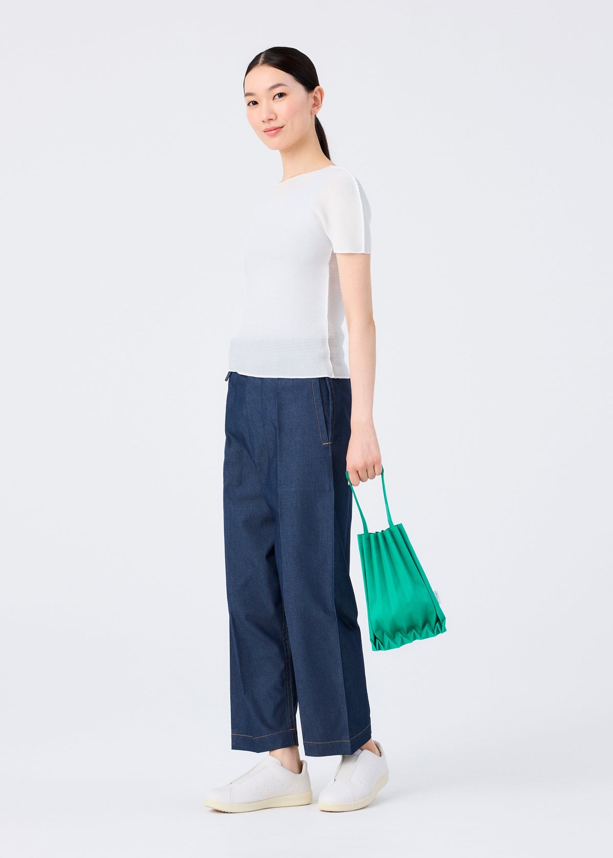 A person stands against a white background wearing a white short-sleeve top, wide-leg blue jeans, white slip-on shoes, and carrying the me ISSEY MIYAKE TRUNK PLEATS BAG 12 as a vivid mini tote. view 7 of 7