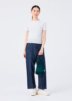 A woman, smiling softly at the camera, stands against a white background wearing a fitted white top, wide-leg blue jeans, white slip-on shoes, and holding the vivid TRUNK PLEATS BAG 12 by me ISSEY MIYAKE.