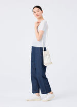 A woman stands sideways in a white short-sleeve top, wide-leg blue jeans, and white sneakers, carrying the vivid TRUNK PLEATS BAG 12 by me ISSEY MIYAKE. She looks over her shoulder with a slight smile against a plain white background.