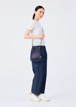 A woman stands sideways, smiling, in a white short-sleeve top, blue wide-leg pants, white slip-on shoes, and carries the me ISSEY MIYAKE TRUNK PLEATS BAG 12 over her shoulder against a plain white background.