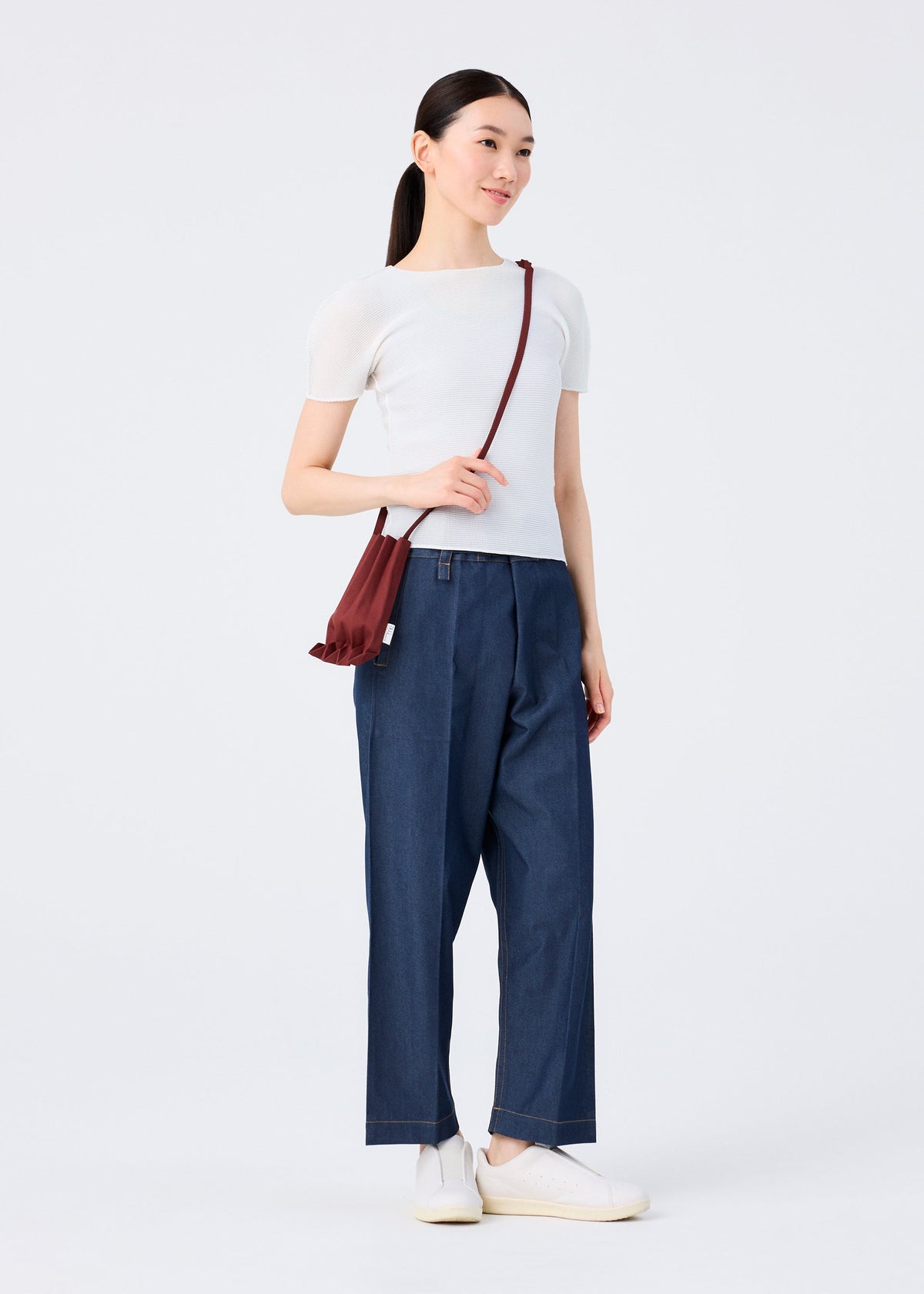 A woman stands against a plain background, smiling and looking aside. She wears a white short-sleeve top, blue wide-leg pants, white slip-on shoes, and carries the me ISSEY MIYAKE TRUNK PLEATS BAG 12 in vivid colors across her shoulder. view 9 of 9