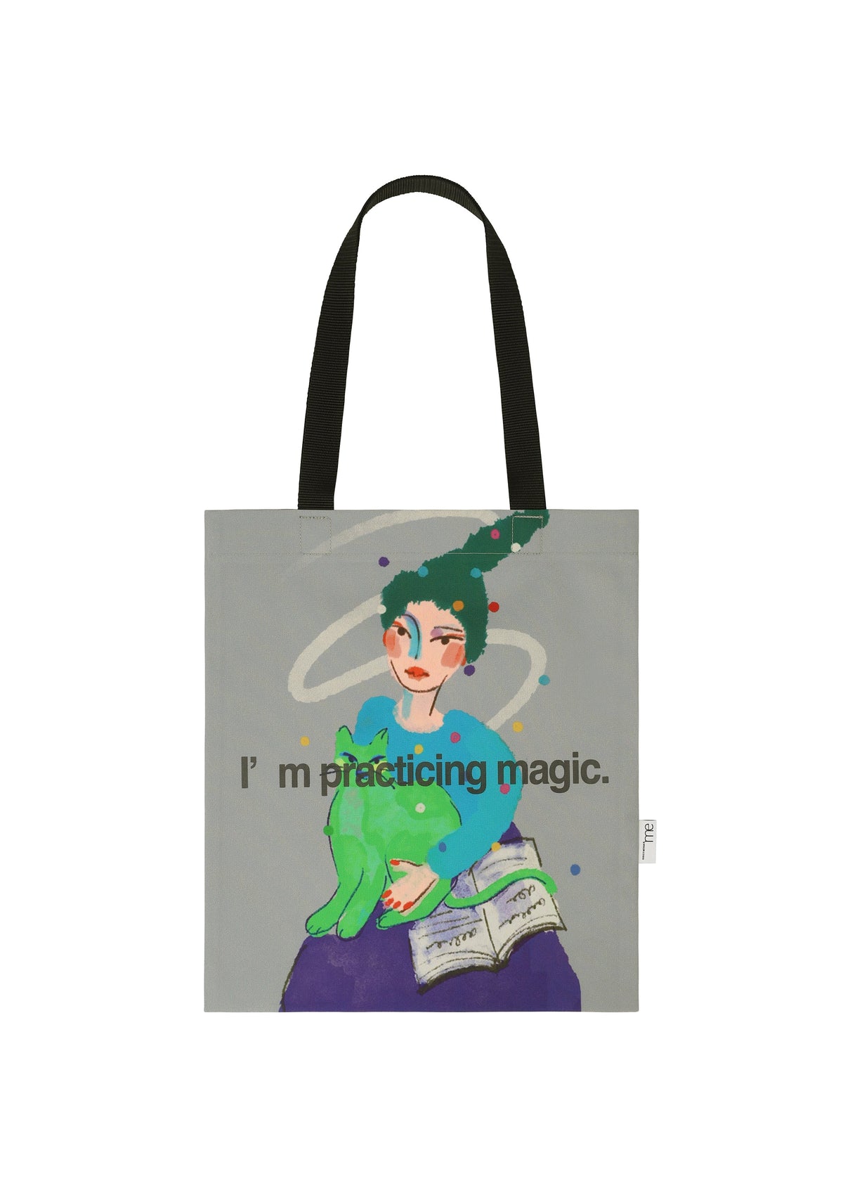 The CAT BAG by me ISSEY MIYAKE is a tote with black handles, showing a person holding a green cat, a floating book, and swirl art. The text says: I’m practicing magic. view 1 of 4