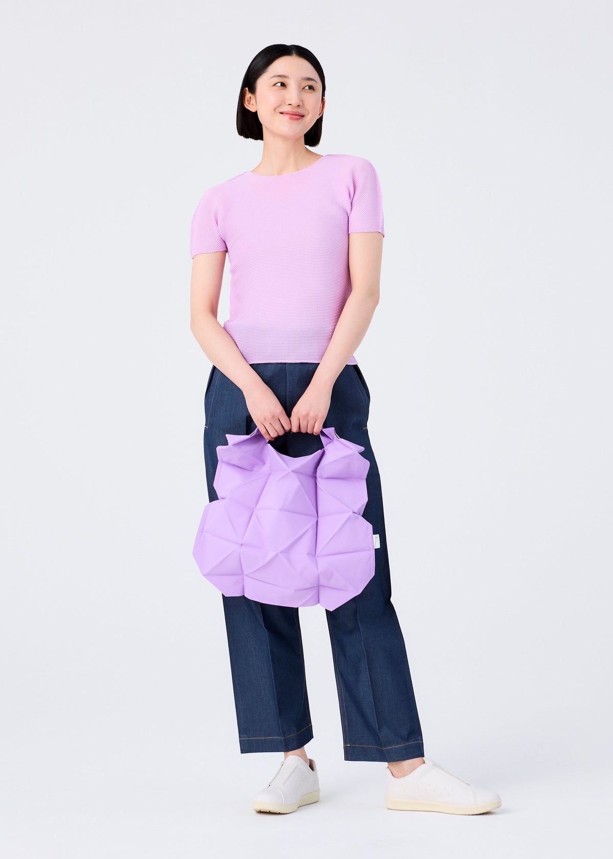 A woman in a light purple top, dark blue jeans, and white shoes holds the GEOMETRIC PLEATS BAG by me ISSEY MIYAKE—a pastel purple bag with handmade pleats and a quadrilateral base—smiling against a plain white background. view 7 of 7