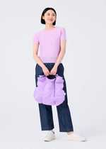 A woman in a light purple top, dark blue jeans, and white shoes holds the GEOMETRIC PLEATS BAG by me ISSEY MIYAKE—a pastel purple bag with handmade pleats and a quadrilateral base—smiling against a plain white background.