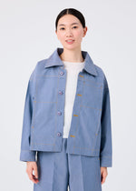 A woman smiles gently with her hands by her sides, wearing the FLAT DENIM light blue button-up jacket and matching wide-leg pants from me ISSEY MIYAKE, crafted from recycled polyester with a realistic denim texture over a white top.