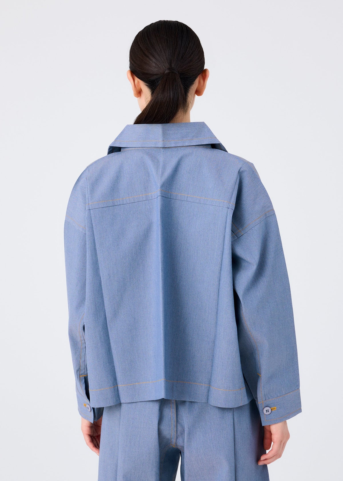 A person with long dark hair in a low ponytail is seen from behind, wearing the FLAT DENIM jacket and pants by me ISSEY MIYAKE, both in recycled polyester with a denim-like texture, against a plain white background. view 6 of 8