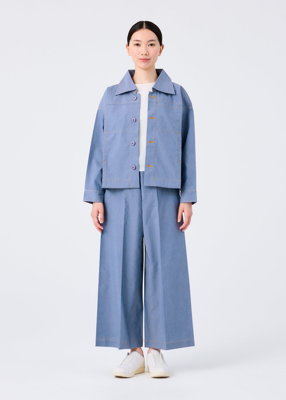 A person stands facing forward in me ISSEY MIYAKE's FLAT DENIM light blue jacket and wide-legged pants made from recycled polyester denim, paired with a white shirt and white slip-on shoes, against a plain white background. view 7 of 8