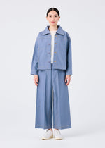 A person stands facing forward in me ISSEY MIYAKE's FLAT DENIM light blue jacket and wide-legged pants made from recycled polyester denim, paired with a white shirt and white slip-on shoes, against a plain white background.