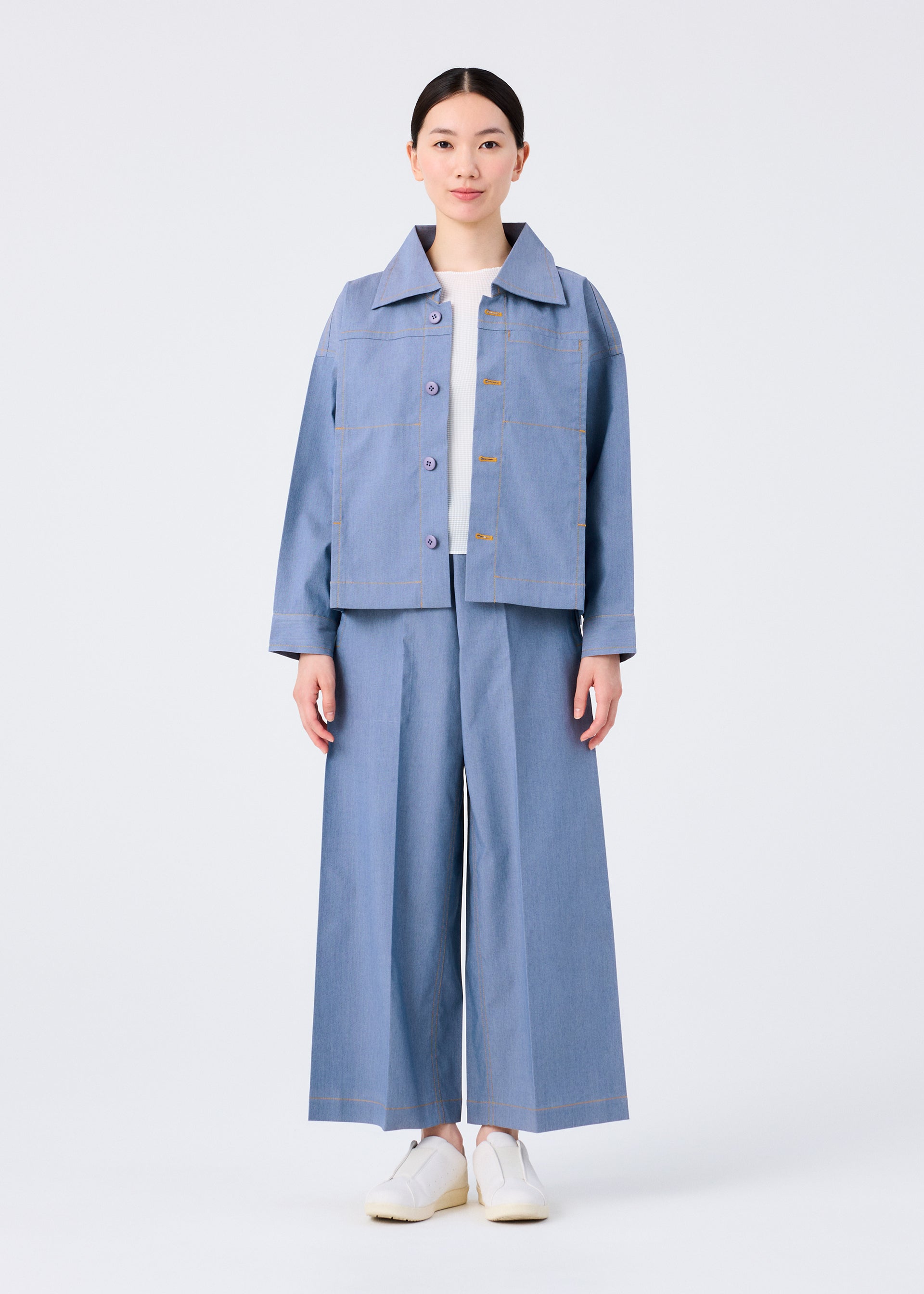 Zoomed view A person stands facing forward in me ISSEY MIYAKE's FLAT DENIM light blue jacket and wide-legged pants made from recycled polyester denim, paired with a white shirt and white slip-on shoes, against a plain white background.