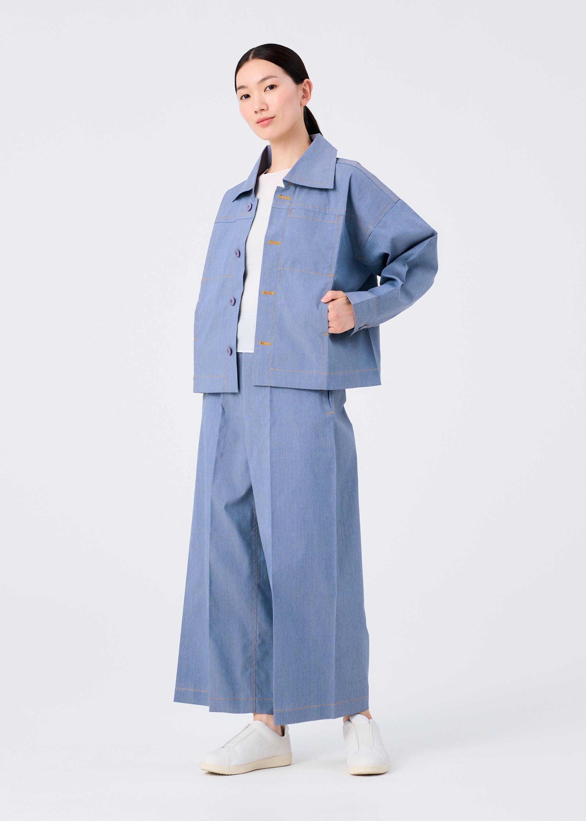 Zoomed view A person stands against a white background wearing the me ISSEY MIYAKE FLAT DENIM jacket and matching wide-leg pants, styled with a white shirt and white slip-on shoes.