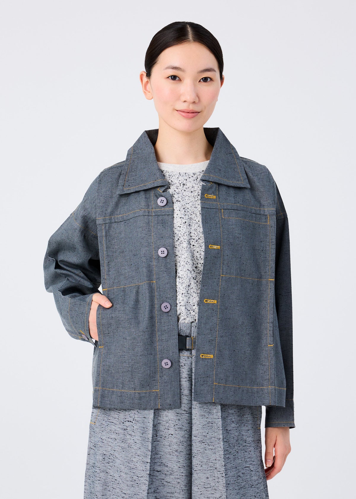 A woman with dark hair pulled back wears the me ISSEY MIYAKE REmeTEX NEP DENIM reversible gray jacket with yellow stitching, made using a wool recycling technique, over a gray patterned dress, standing against a plain light background. view 8 of 12