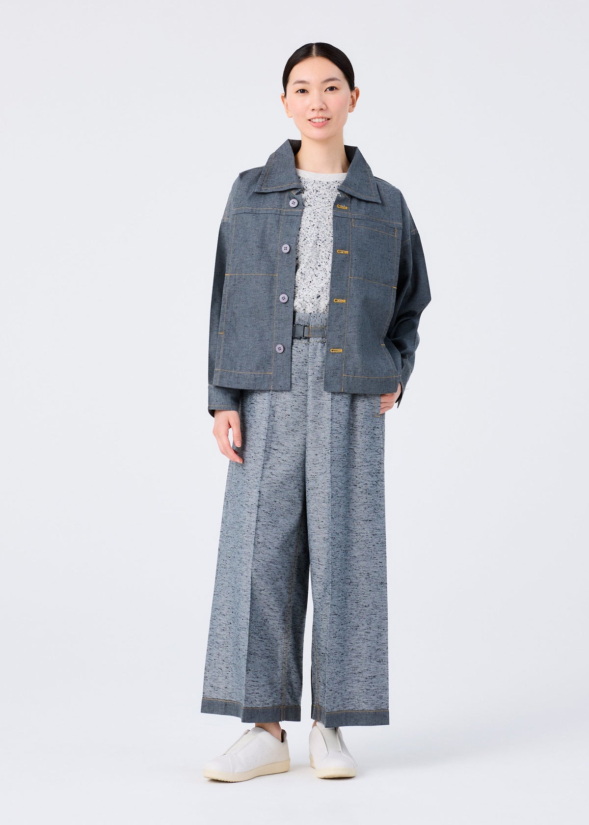 A person faces forward in a me ISSEY MIYAKE REmeTEX NEP DENIM blue jacket, wide-leg gray trousers, textured white top, and white shoes against a plain white background. view 10 of 12