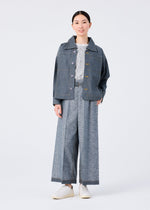 A person faces forward in a me ISSEY MIYAKE REmeTEX NEP DENIM blue jacket, wide-leg gray trousers, textured white top, and white shoes against a plain white background.