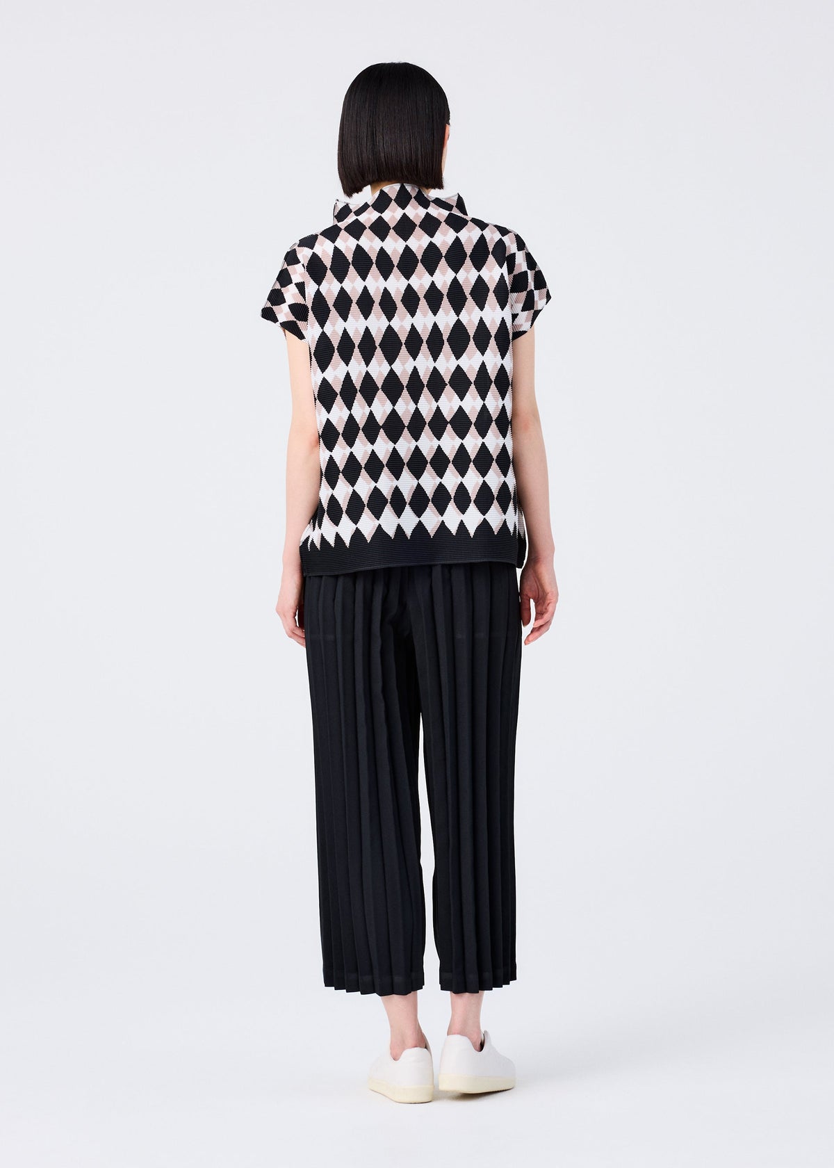 A person stands facing away, wearing me ISSEY MIYAKE COLOR PLEATS BOTTOM pants with a short-sleeve, black-and-white diamond-patterned top and white slip-on shoes against a plain light background. view 6 of 6