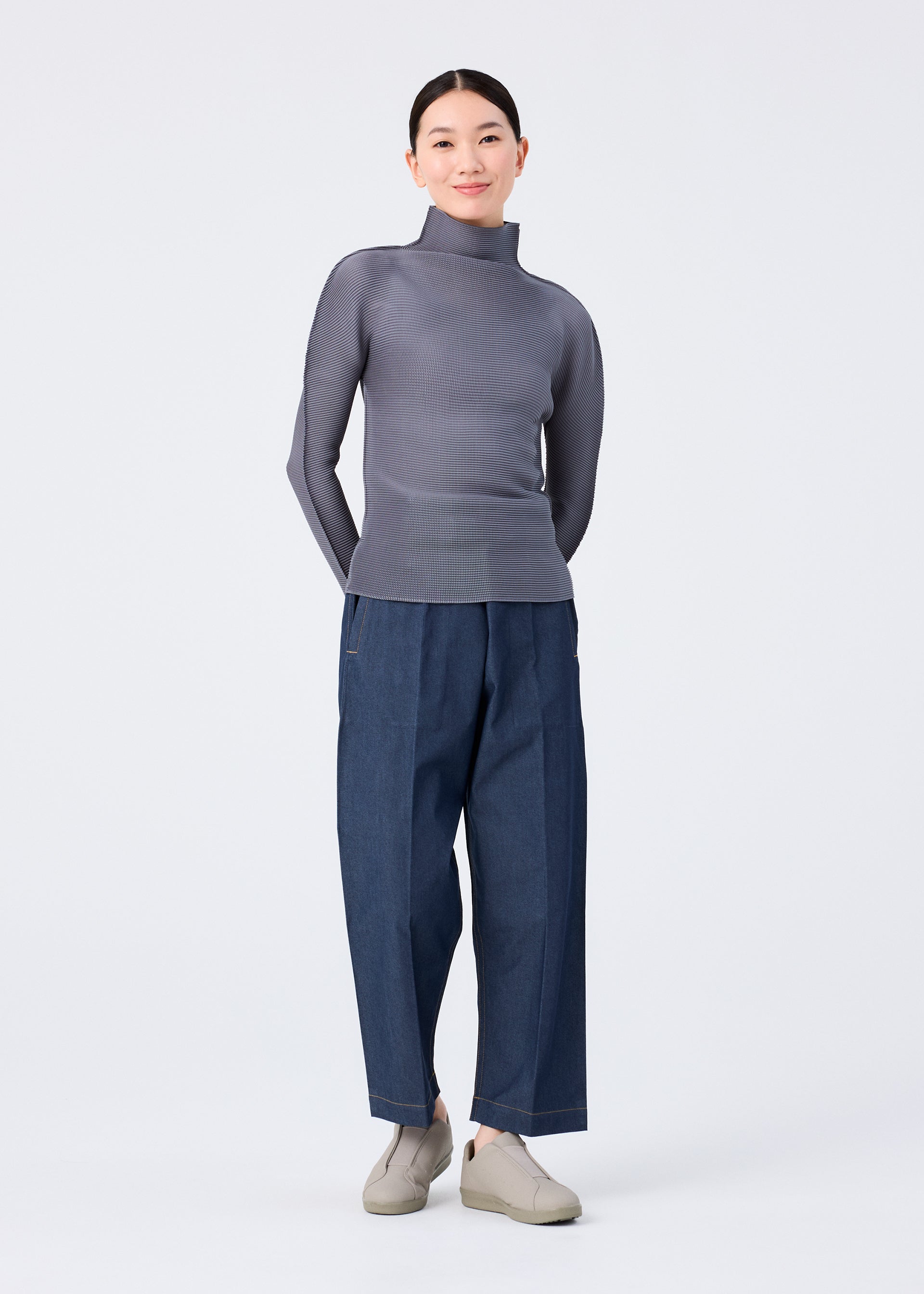 Zoomed view A woman stands against a plain white background, softly smiling with her hands behind her back. She wears the FLAT DENIM pants by me ISSEY MIYAKE, featuring a realistic denim texture made from recycled polyester denim and styled with a gray turtleneck sweater.