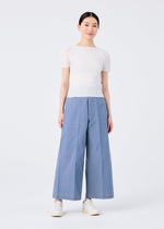 A woman stands against a white background, wearing a fitted short-sleeve white top, me ISSEY MIYAKE FLAT DENIM wide pants made from recycled polyester denim, and white slip-on shoes, smiling slightly with her hands at her sides.