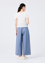 A woman with long dark hair in a ponytail stands with her back to the camera, wearing a short-sleeved white top and me ISSEY MIYAKE FLAT DENIM wide pants made from recycled polyester denim, plus white slip-on shoes, against a plain background.