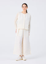 A woman smiles against a white backdrop, dressed in the me ISSEY MIYAKE RIPPLE PLEATS textured white vest with handmade pleats and wide matching pocketed pants, layered over a beige long-sleeve top and white shoes.