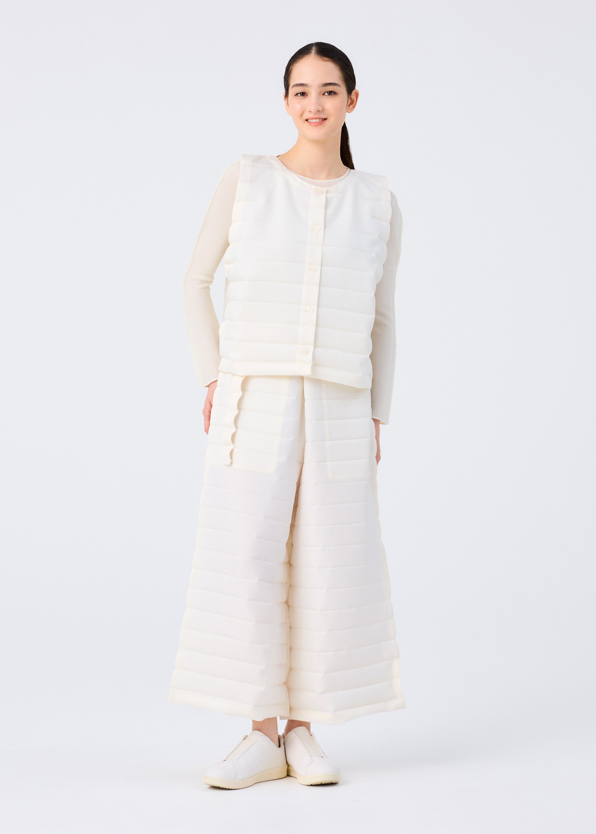 Zoomed view A woman smiles against a white backdrop, dressed in the me ISSEY MIYAKE RIPPLE PLEATS textured white vest with handmade pleats and wide matching pocketed pants, layered over a beige long-sleeve top and white shoes.