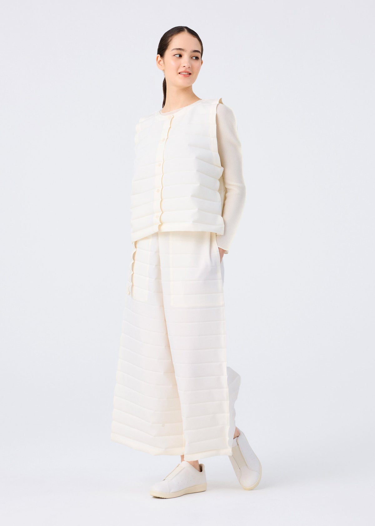 A woman models the me ISSEY MIYAKE RIPPLE PLEATS set—an off-white textured long-sleeve top and wide pants with handmade pleats and discreet pockets—paired with white slip-on shoes, standing against a plain white background. view 7 of 7