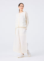 A woman models the me ISSEY MIYAKE RIPPLE PLEATS set—an off-white textured long-sleeve top and wide pants with handmade pleats and discreet pockets—paired with white slip-on shoes, standing against a plain white background.
