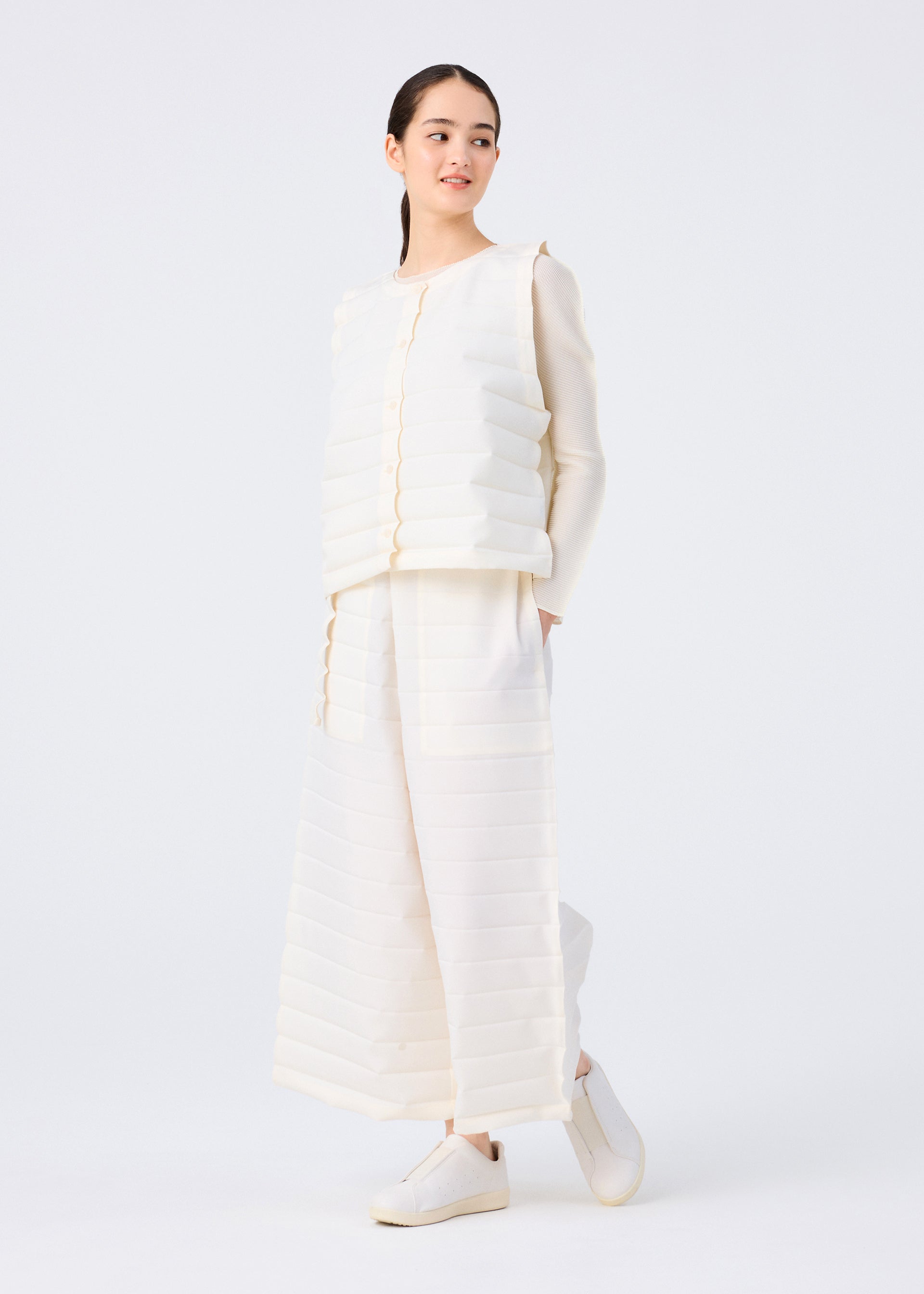 Zoomed view A woman models the me ISSEY MIYAKE RIPPLE PLEATS set—an off-white textured long-sleeve top and wide pants with handmade pleats and discreet pockets—paired with white slip-on shoes, standing against a plain white background.