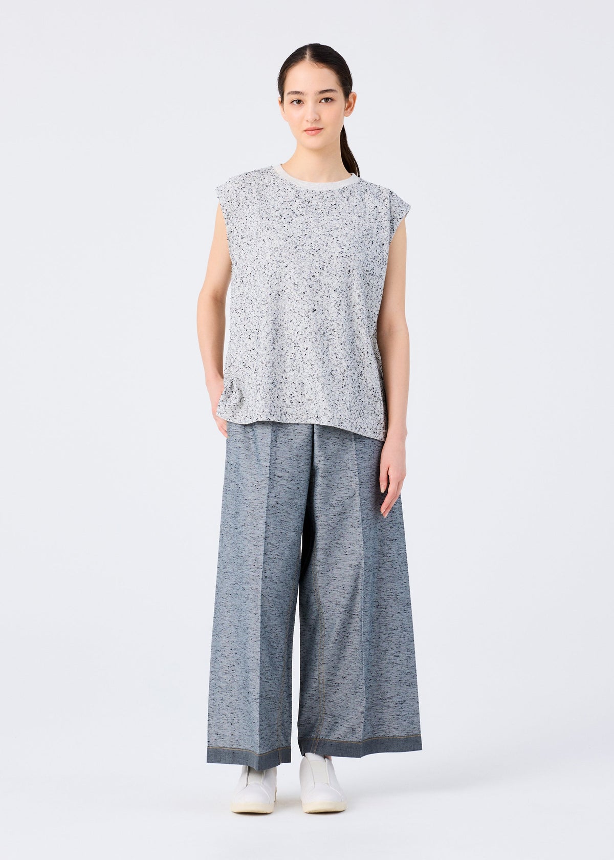 A person with long dark hair tied back and a neutral expression stands against a plain white background, wearing wide-legged gray REmeTEX NEP DENIM pants by me ISSEY MIYAKE, a loose patterned top, and white shoes. view 5 of 6