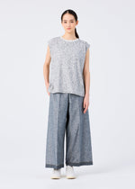 A person with long dark hair tied back and a neutral expression stands against a plain white background, wearing wide-legged gray REmeTEX NEP DENIM pants by me ISSEY MIYAKE, a loose patterned top, and white shoes.