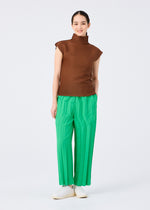 A person stands against a white background, smiling with hands in their pockets. They wear a sleeveless brown turtleneck top, me ISSEY MIYAKE's ZIG ZAG PLEATS bright green pants, and white slip-on shoes.