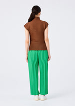 A woman stands facing away from the camera, wearing a short-sleeved brown top and bright green me ISSEY MIYAKE ZIG ZAG PLEATS pants with a zigzag silhouette, along with white shoes, set against a plain white background.