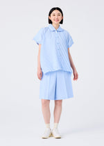 A person with short dark hair smiles while wearing the FLAT BOTTOM set by me ISSEY MIYAKE: a light blue short-sleeve button-up shirt and matching pressed shorts, paired with white socks and slip-on shoes against a plain white background.