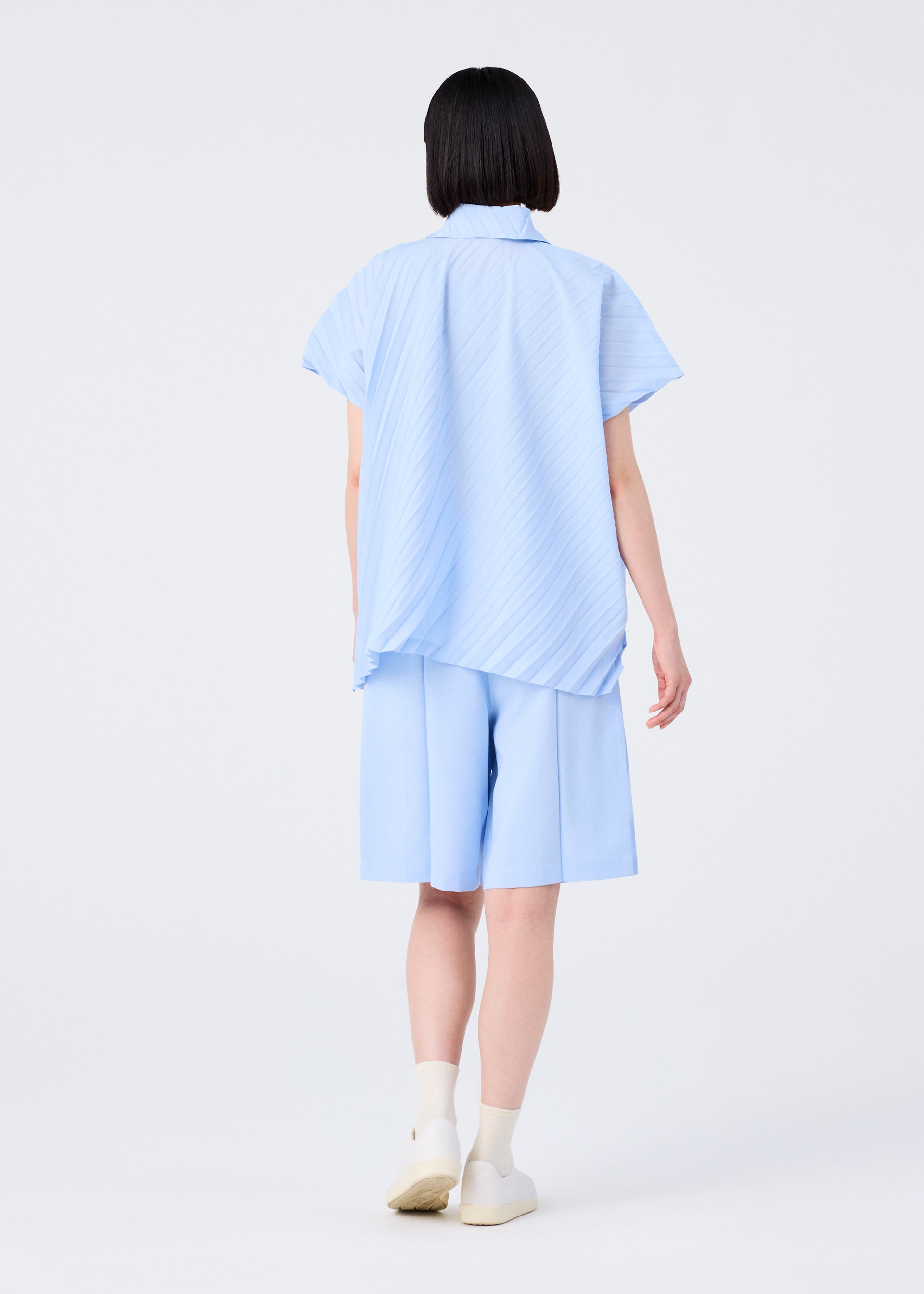 Zoomed view A person with short black hair, seen from behind, wears me ISSEY MIYAKE's FLAT BOTTOM pressed knee-length shorts with a subtle melange effect, paired with a light blue shirt, white socks, and white shoes against a plain light background.