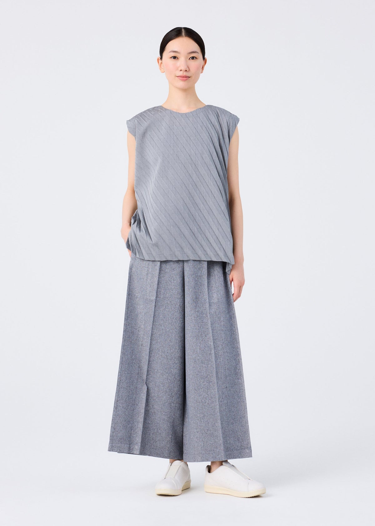 A person stands against a white background wearing me ISSEY MIYAKE's FLAT BOTTOM: sleeveless light gray pleated top and pressed wide pants with a flowy feel, paired with white slip-on shoes. Hair is pulled back, arms relaxed by their sides. view 7 of 8