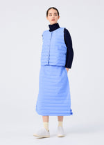 A woman stands against a white background wearing the RIPPLE PLEATS sleeveless light blue vest and matching skirt with pockets by me ISSEY MIYAKE over a black turtleneck, paired with white socks and slip-on shoes, gazing calmly to the side.