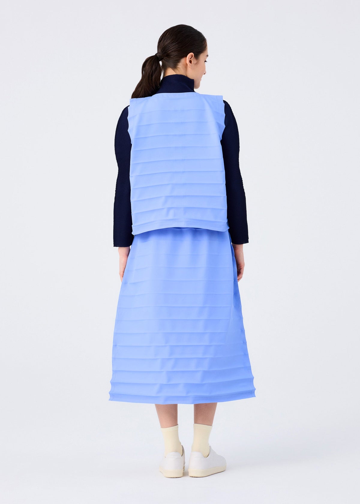 A woman with tied-back dark hair stands facing away, wearing the me ISSEY MIYAKE RIPPLE PLEATS light blue midi skirt and matching vest over a black long-sleeve top and white shoes, set against a plain white background. view 7 of 7