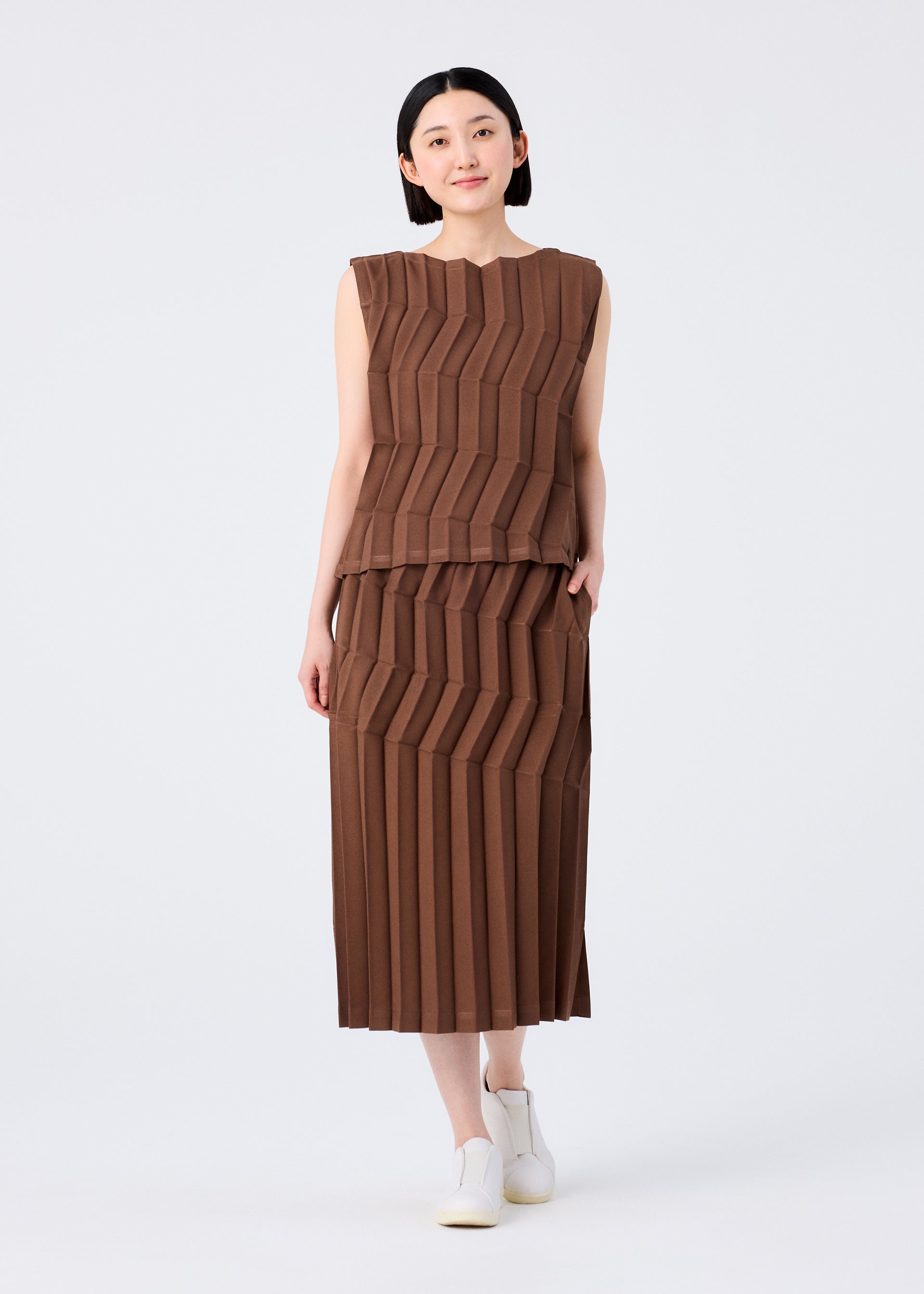 Zoomed view A woman with short black hair smiles softly, wearing the ZIG ZAG PLEATS sleeveless top and hand-pleated skirt by me ISSEY MIYAKE against a plain background. She pairs the geometric-patterned brown set with white shoes, one hand in her pocket.