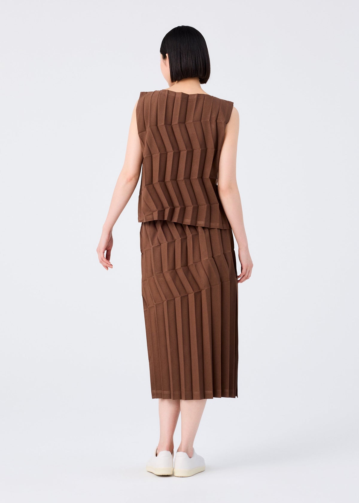 A person with short dark hair, seen from behind, wears the ZIG ZAG PLEATS top and matching skirt by me ISSEY MIYAKE. The brown, sleeveless set features a zigzag silhouette. They stand against a plain white background in white shoes. view 6 of 7