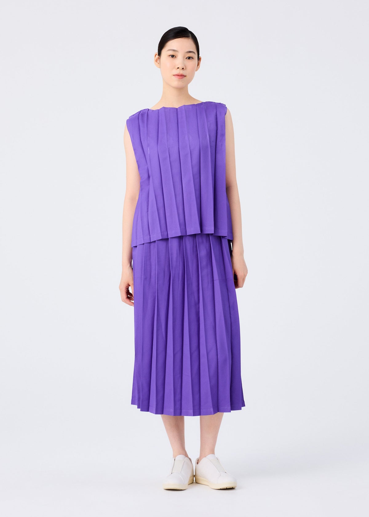 A woman faces forward against a white background, wearing the me ISSEY MIYAKE CROSS PLEATS sleeveless purple top and matching hand-pleated skirt, paired with white slip-on shoes. view 6 of 7