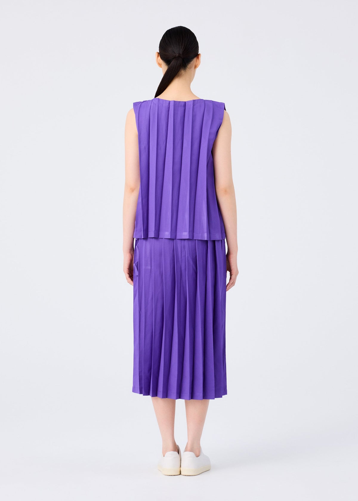 A person with long dark hair in a low ponytail is seen from behind, wearing the CROSS PLEATS sleeveless purple top and hand-pleated skirt by me ISSEY MIYAKE, paired with white slip-on shoes against a plain white background. view 7 of 7
