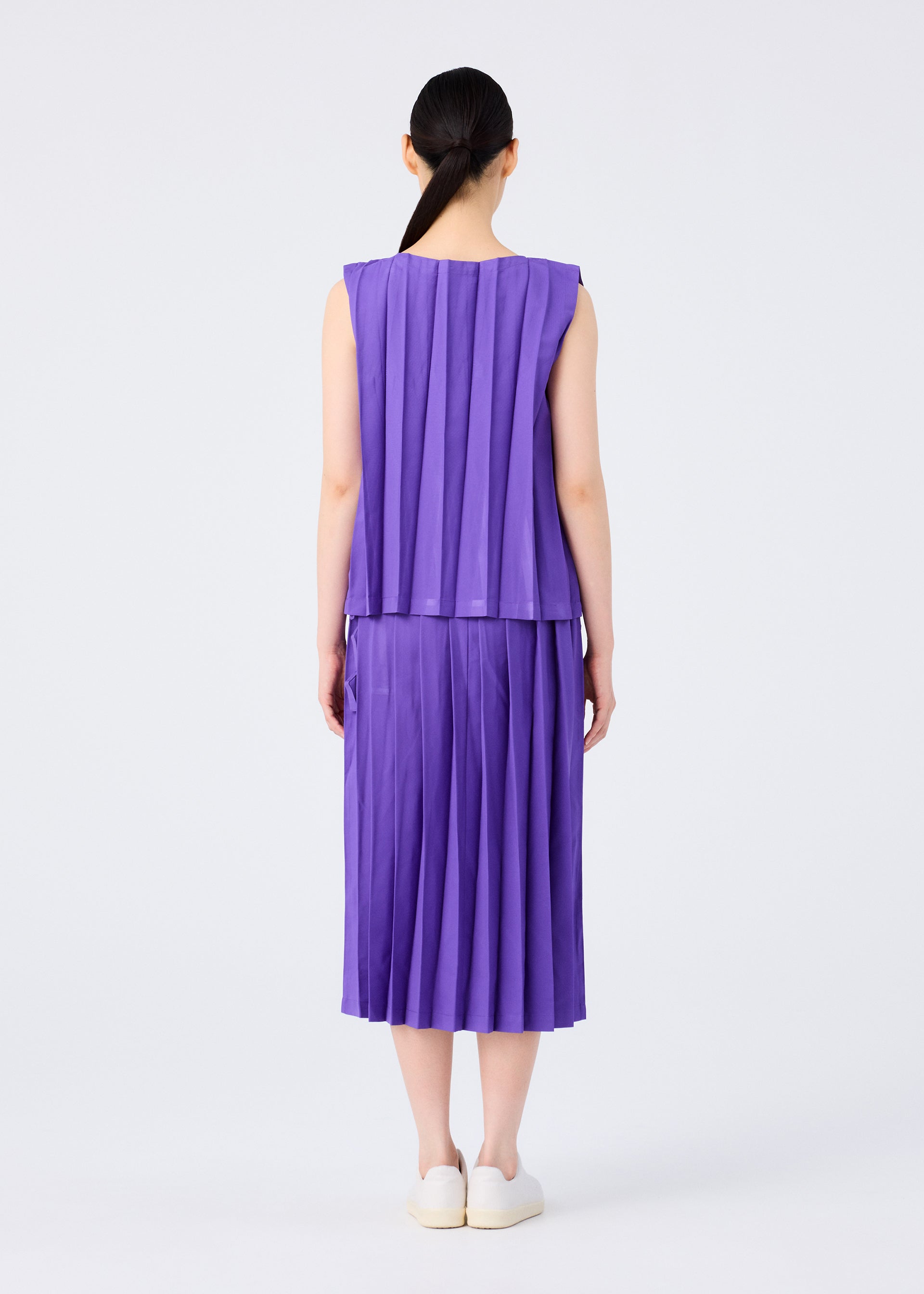 Zoomed view A person with long dark hair in a low ponytail is seen from behind, wearing the CROSS PLEATS sleeveless purple top and hand-pleated skirt by me ISSEY MIYAKE, paired with white slip-on shoes against a plain white background.