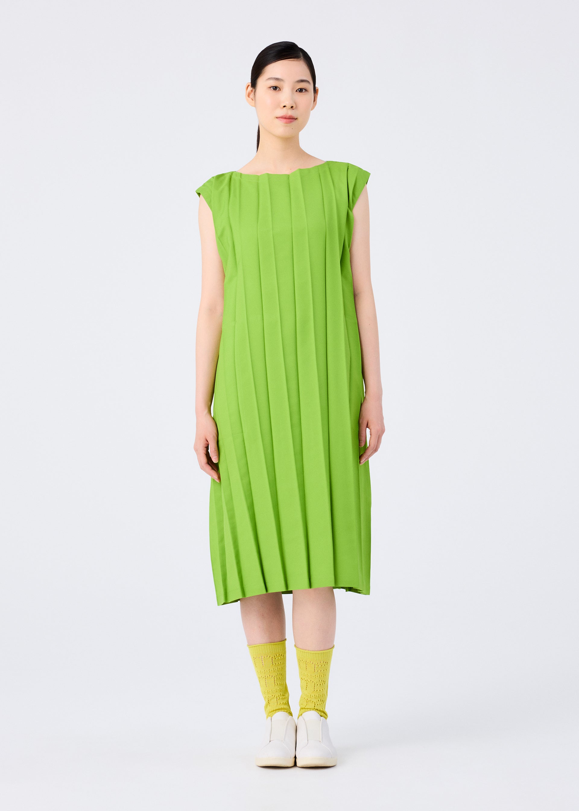 Zoomed view A woman faces forward in the CROSS PLEATS knee-length dress by me ISSEY MIYAKE, featuring bright green hand-pleating and cap sleeves. She pairs it with yellow socks and white shoes against a plain, light background.