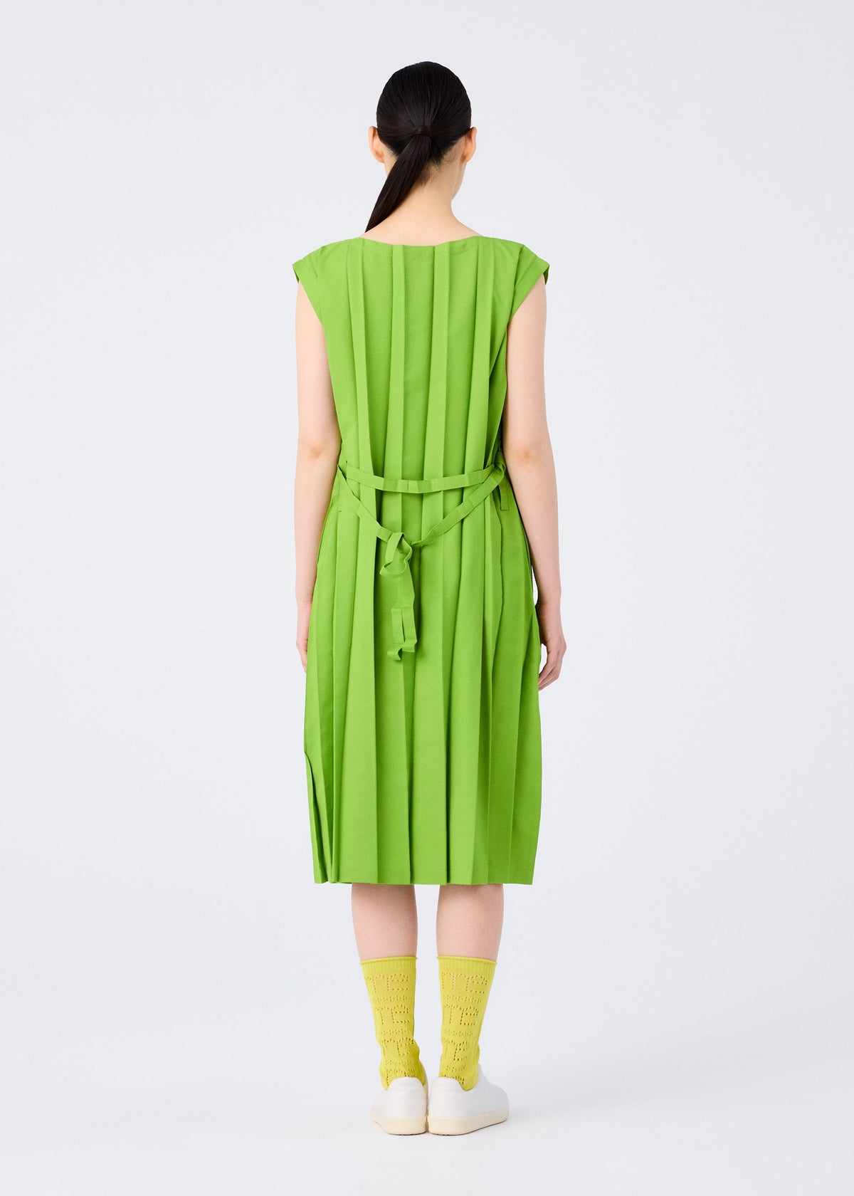 A woman faces away, wearing the me ISSEY MIYAKE CROSS PLEATS bright green hand-pleated dress with a back-tied belt. She has straight dark hair in a low ponytail, yellow socks, and white shoes against a plain light background. view 7 of 8