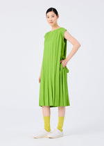 A woman stands against a white background, smiling with one hand on her hip, wearing the CROSS PLEATS sleeveless dress in bright green by me ISSEY MIYAKE, paired with yellow textured socks and white slip-on shoes.