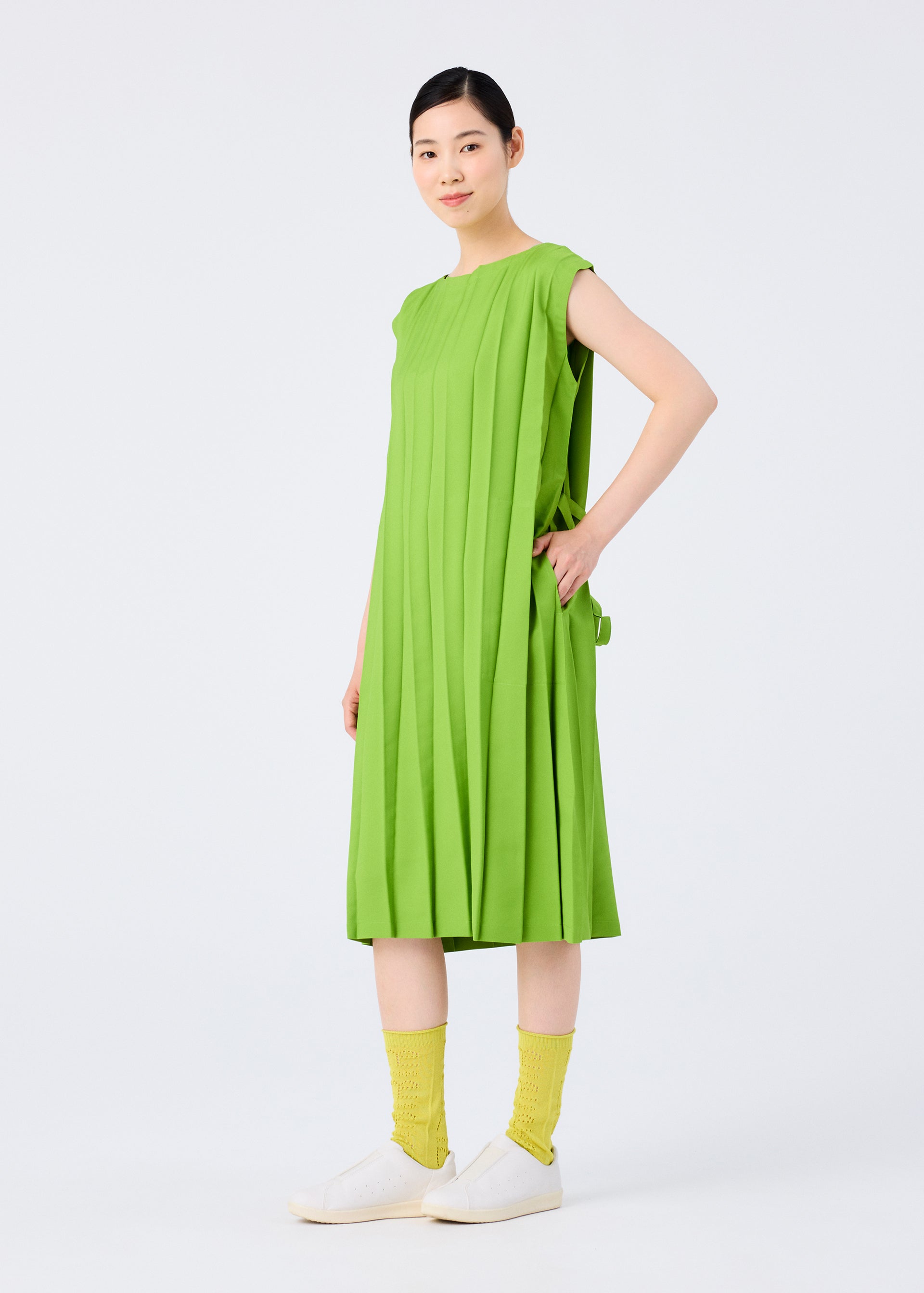 Zoomed view A woman stands against a white background, smiling with one hand on her hip, wearing the CROSS PLEATS sleeveless dress in bright green by me ISSEY MIYAKE, paired with yellow textured socks and white slip-on shoes.