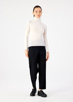 A person with dark hair tied back and a neutral expression stands against a white background, wearing a white high-neck top, black STRETCH PLEATS pants by me ISSEY MIYAKE, and black slip-on shoes.
